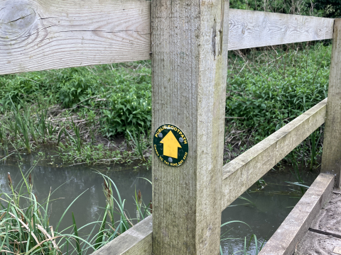 Photograph of a footbridge across a chalk stream with a public footpath sign attached