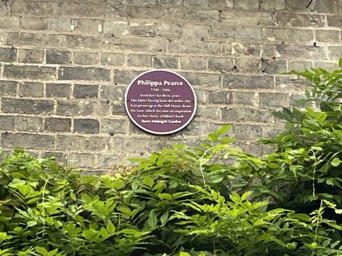 A photograph of the dark red plaque to Philippa Pearce. The inscription reads "Her father having been the miller, she had grown up in the Mill House down the lane, which became an inspiration for her classic children's book Tom's Midnight Garden."