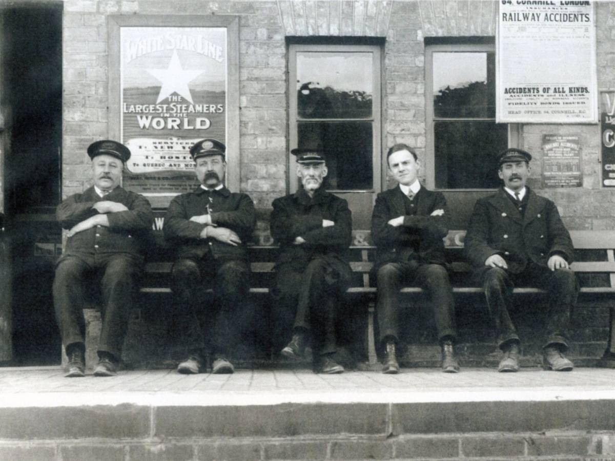 Black and white photograph of four men sitting on a bench outside Meldreth railway station