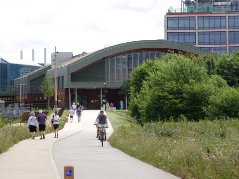 A photograph taken from Hobson's Park showing the entrance to Cambridge South Station. There are people on the footpaths and cyclists on the cycle path.